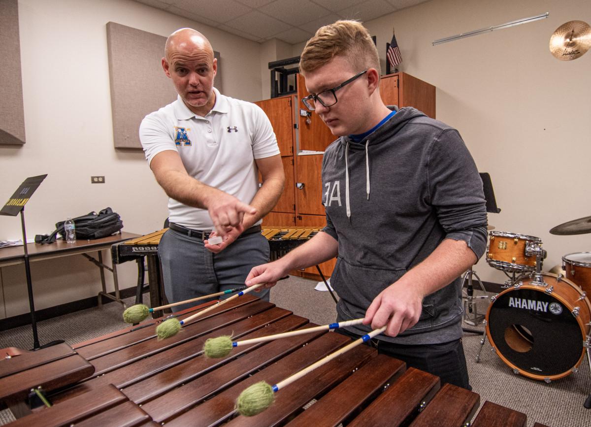 Student plays on percussion instrument with band director in background.